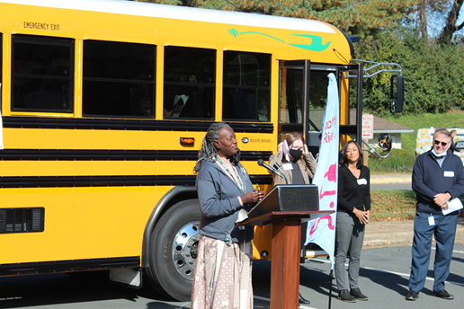 Louisa county's Deborah Coles stands at a podium in front to an electric school bus in Charlottesville Virginia. She is smiling as she share's her county's experience with their electric school buses.